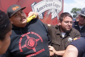 A cop holds a taser on a man's neck as he stands behind a Budweiser truck.